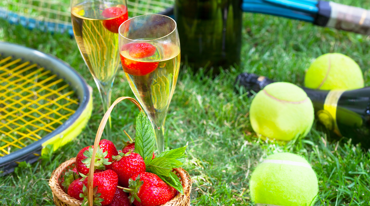 Raquettes et balles de tennis accompagnés de deux coupes de champagnes et un panier rempli de fraises sur du gazon.
